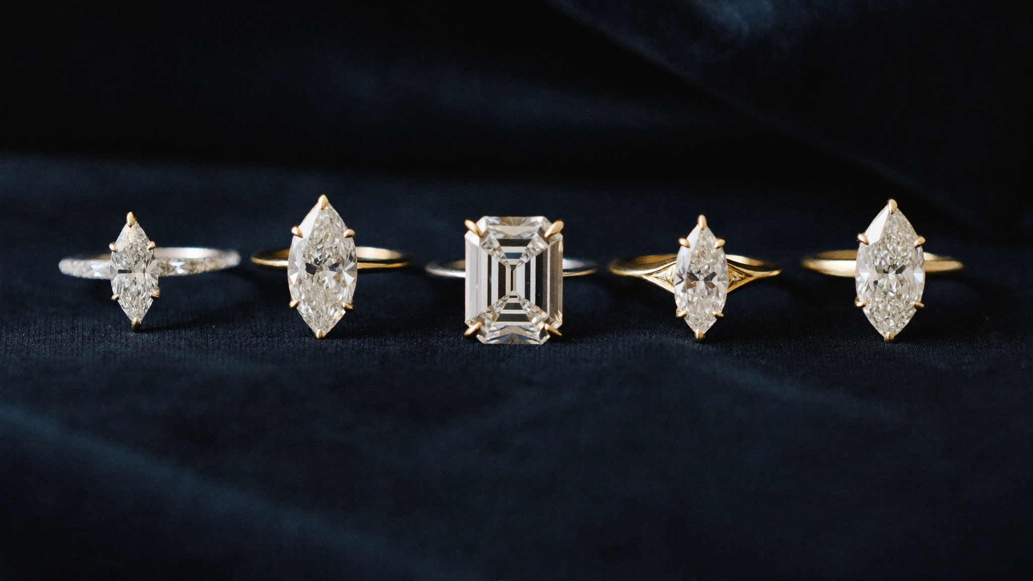 Five diamond cuts — round, oval, emerald, pear, and marquise — arranged on dark velvet under soft directional light