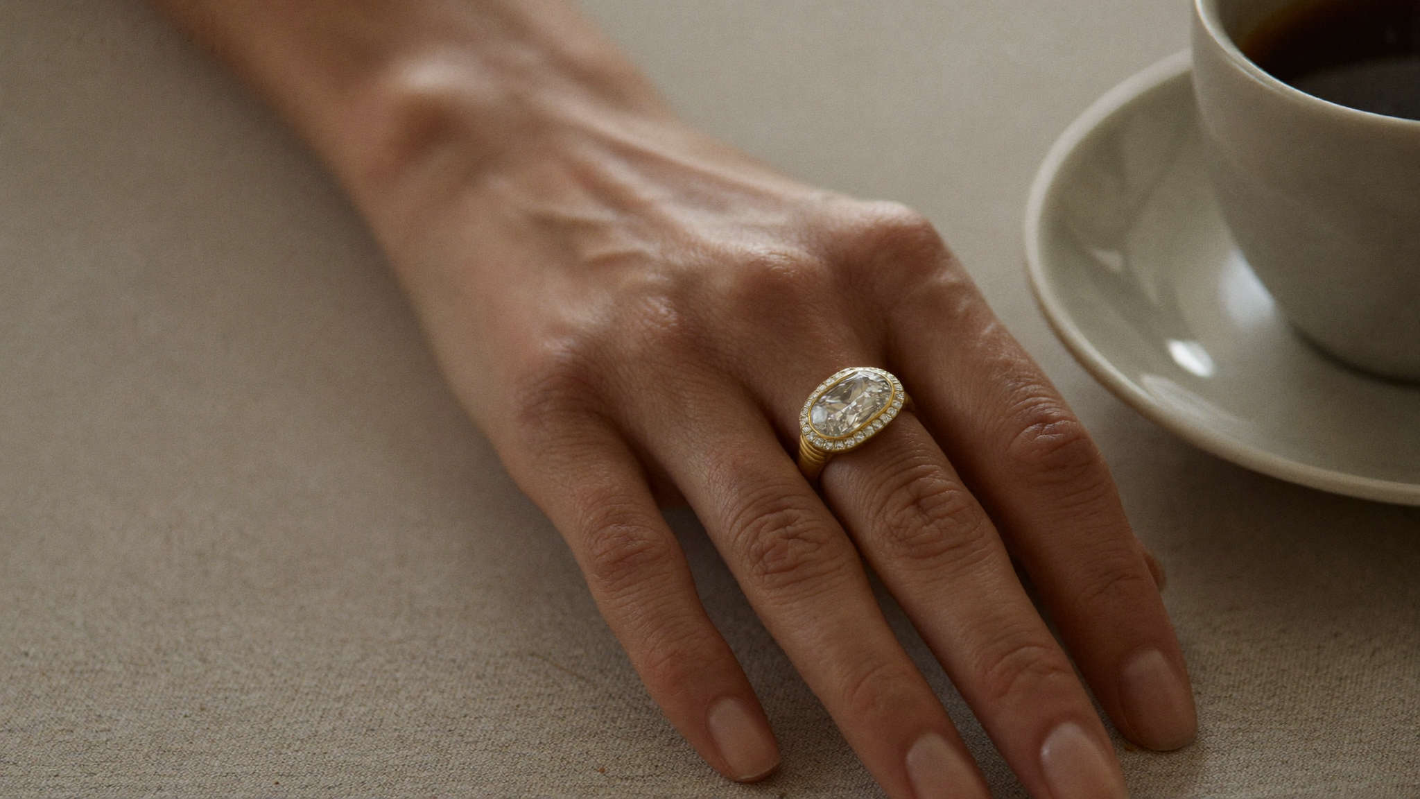 A hand wearing a sleek bezel-set gold diamond ring resting on a linen-draped surface in warm natural light