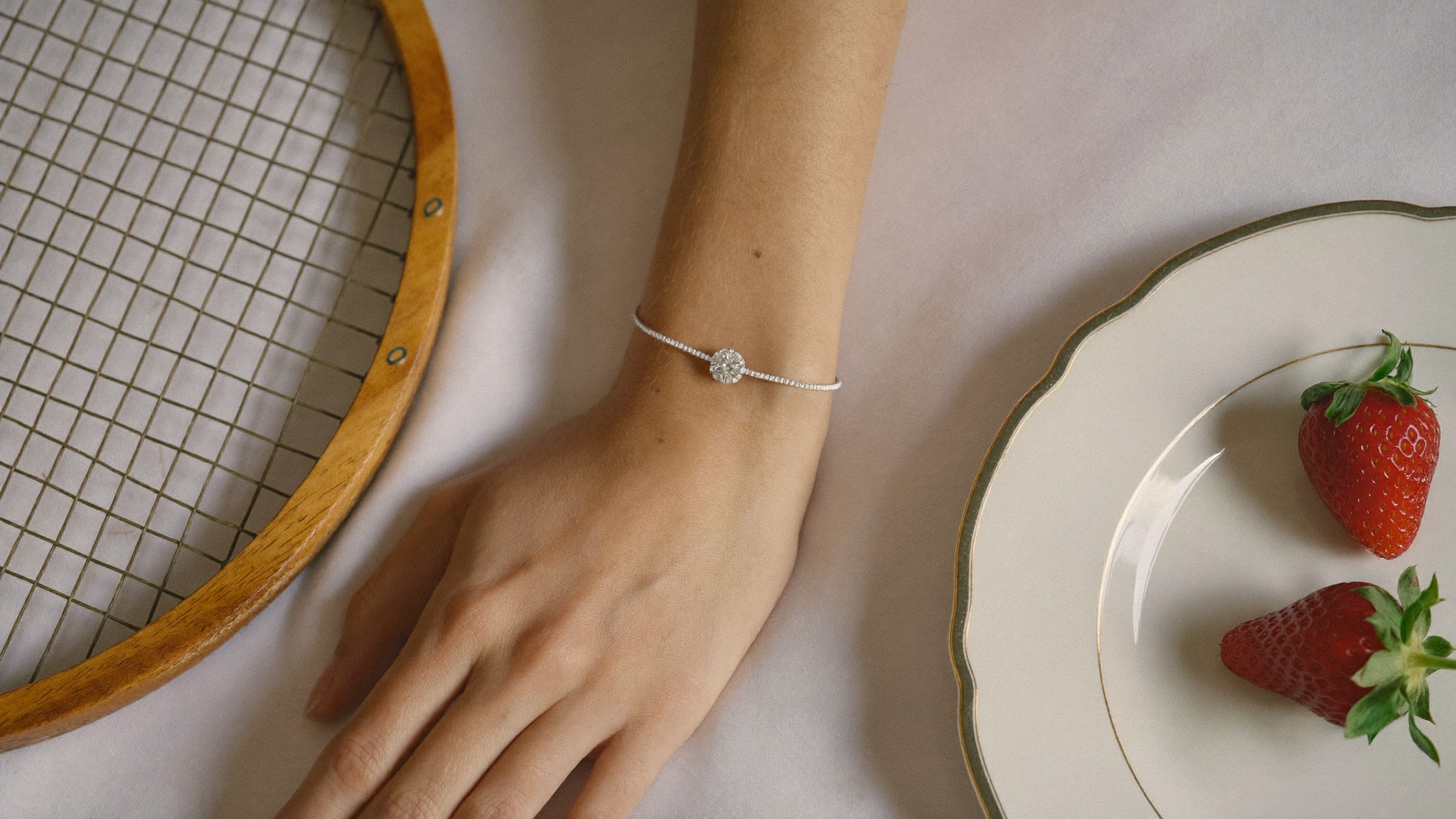 Lab grown diamond tennis bracelet on a woman's wrist beside a vintage tennis racquet on white linen, soft natural light