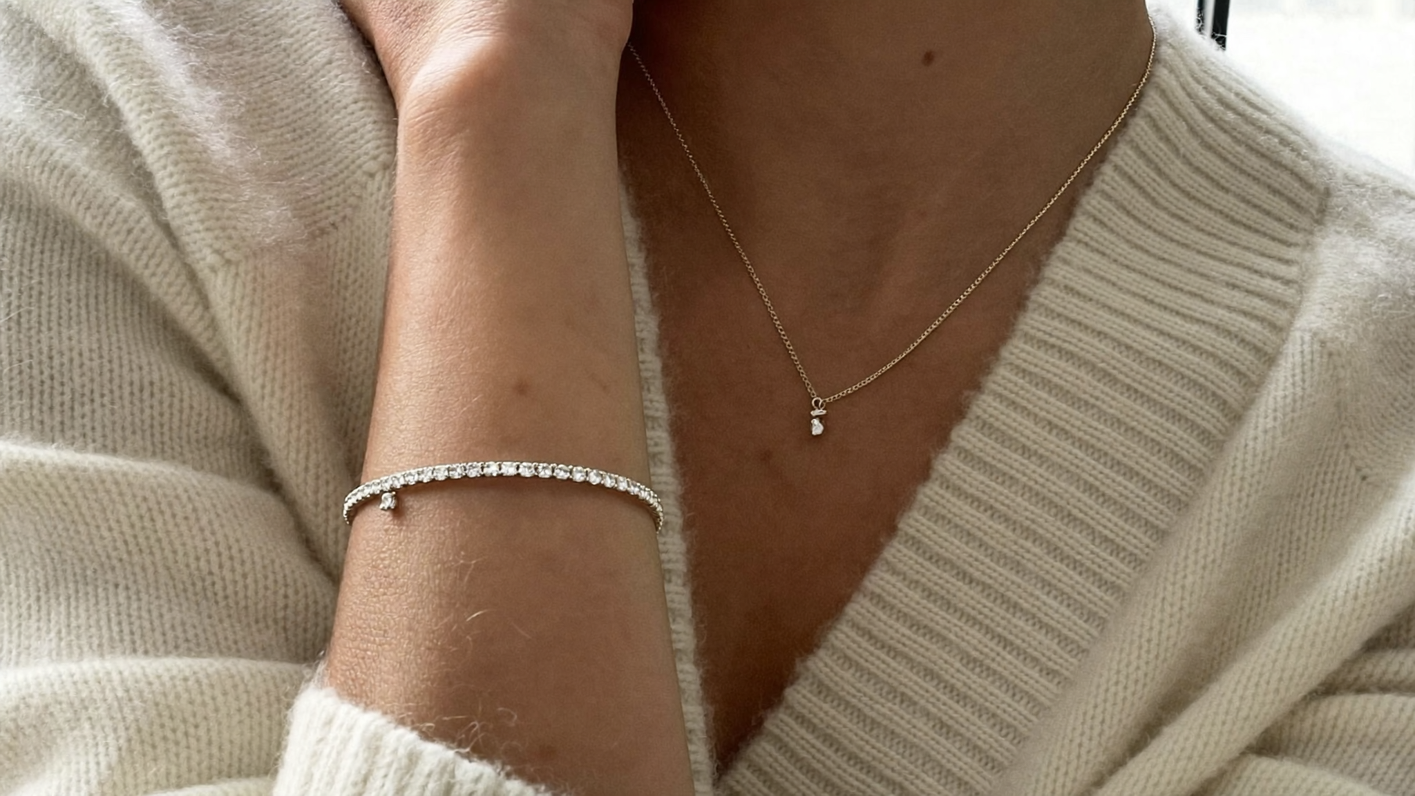 Quiet luxury jewelry styled on a woman's wrist and collarbone: a slim gold diamond tennis bracelet, thin eternity band, and tiny pendant over cream cashmere