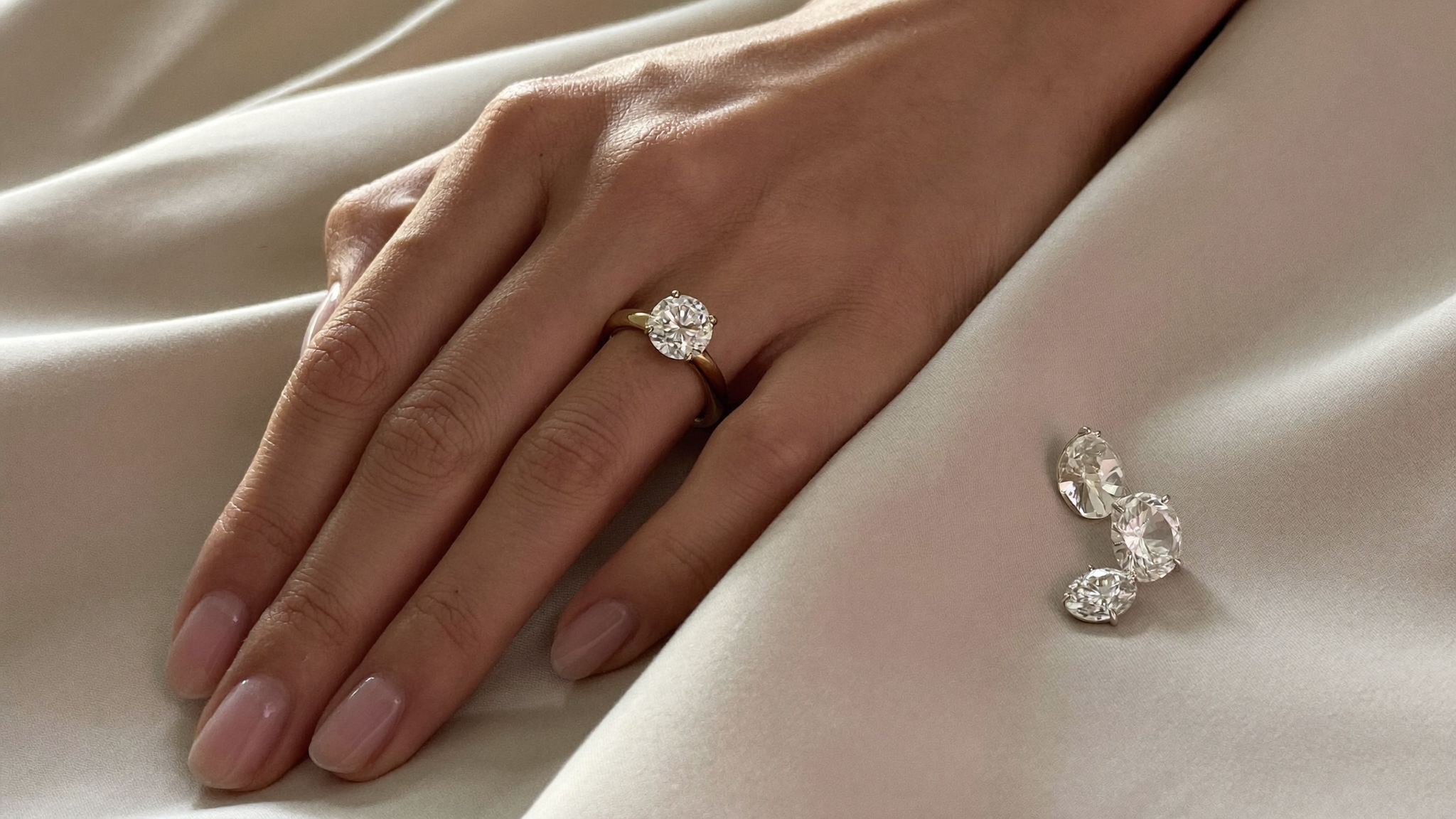 Hand wearing a round lab-grown diamond solitaire ring beside loose diamonds on silk, showing diamond carat size