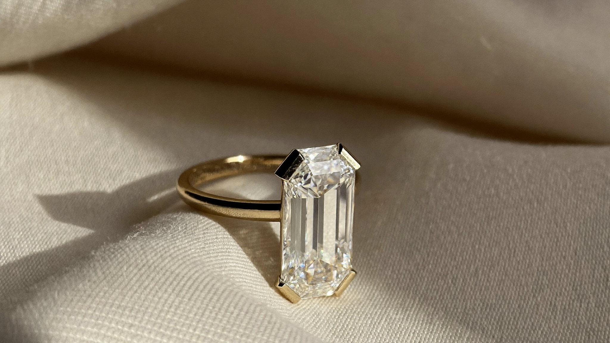 Elongated radiant cut diamond engagement ring in solid yellow gold resting on cream linen in natural light