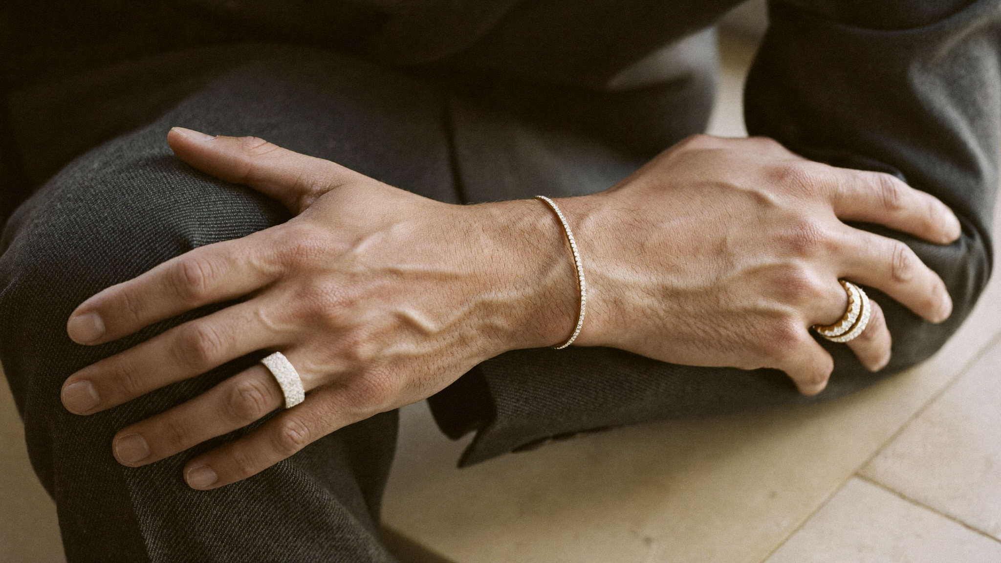Man's wrist wearing a slim gold diamond tennis bracelet and an architectural diamond band, men's diamond jewelry styled with charcoal wool