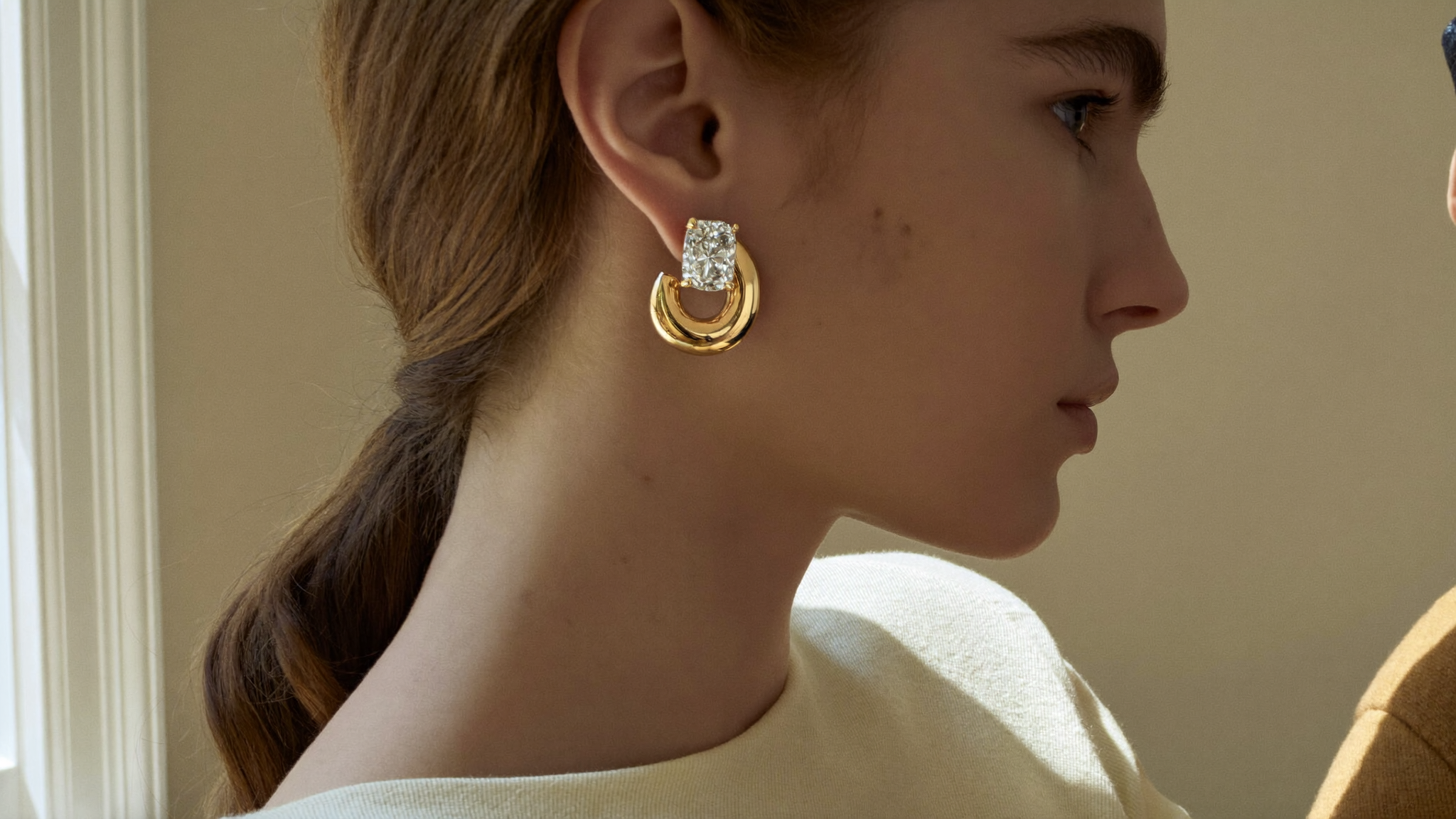 Close-up profile of a woman wearing sculptural solid gold diamond hoop earrings in soft natural window light
