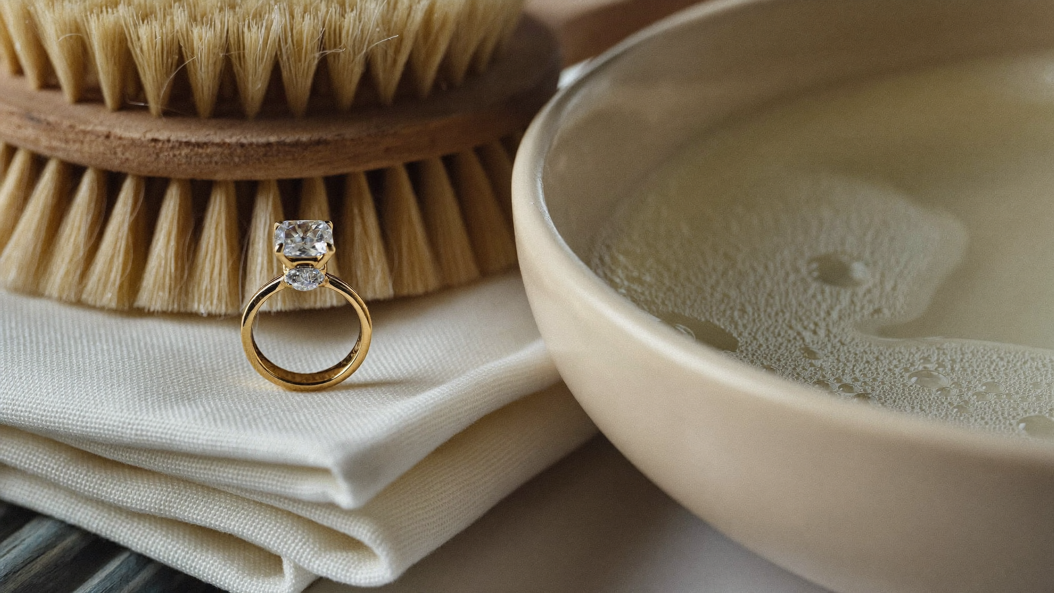 How to clean a diamond ring at home: a solid gold diamond ring on linen beside a soft brush and a bowl of warm soapy water