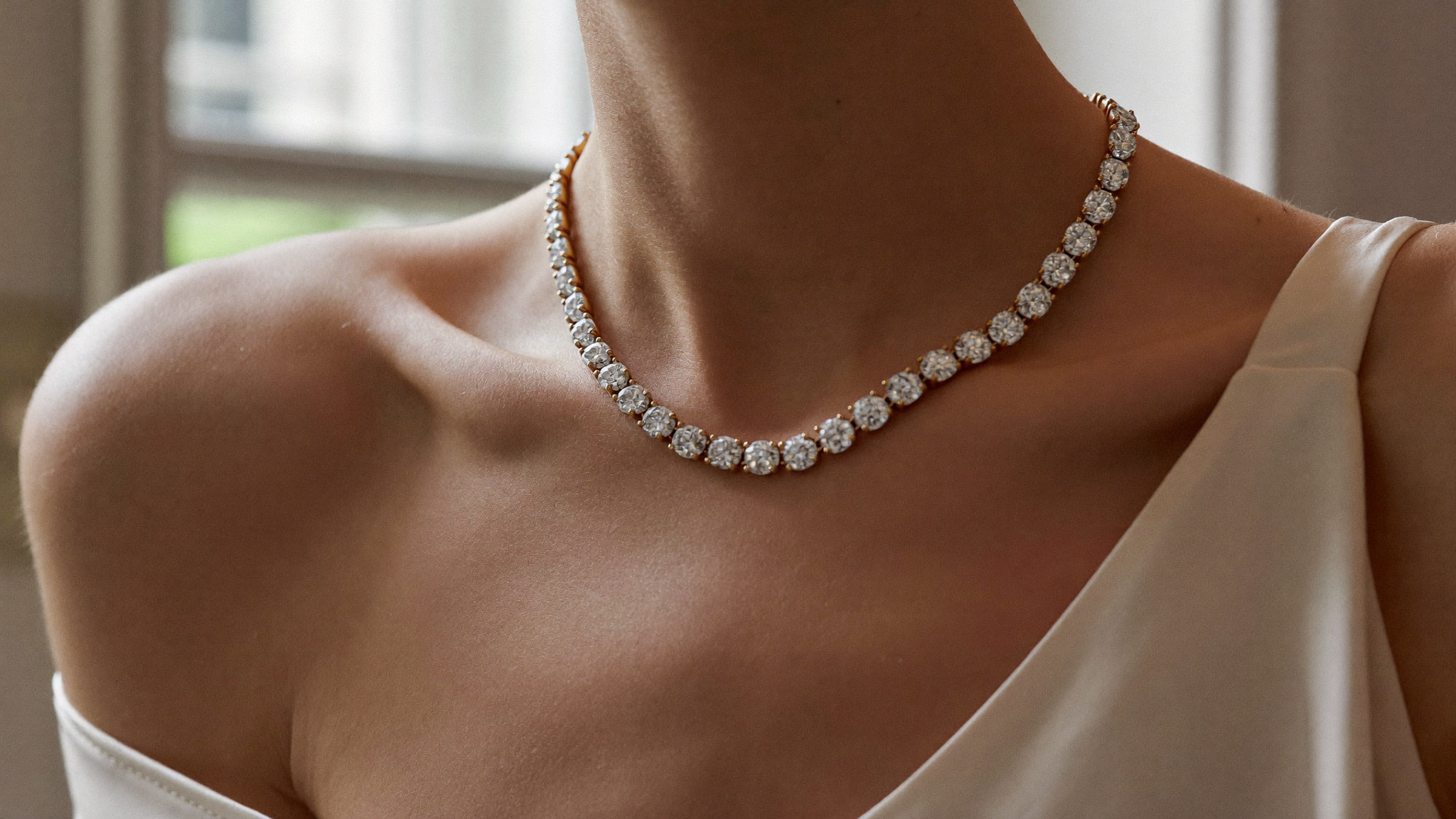 Diamond tennis necklace in gold worn across the collarbone with a cream silk slip, natural window light