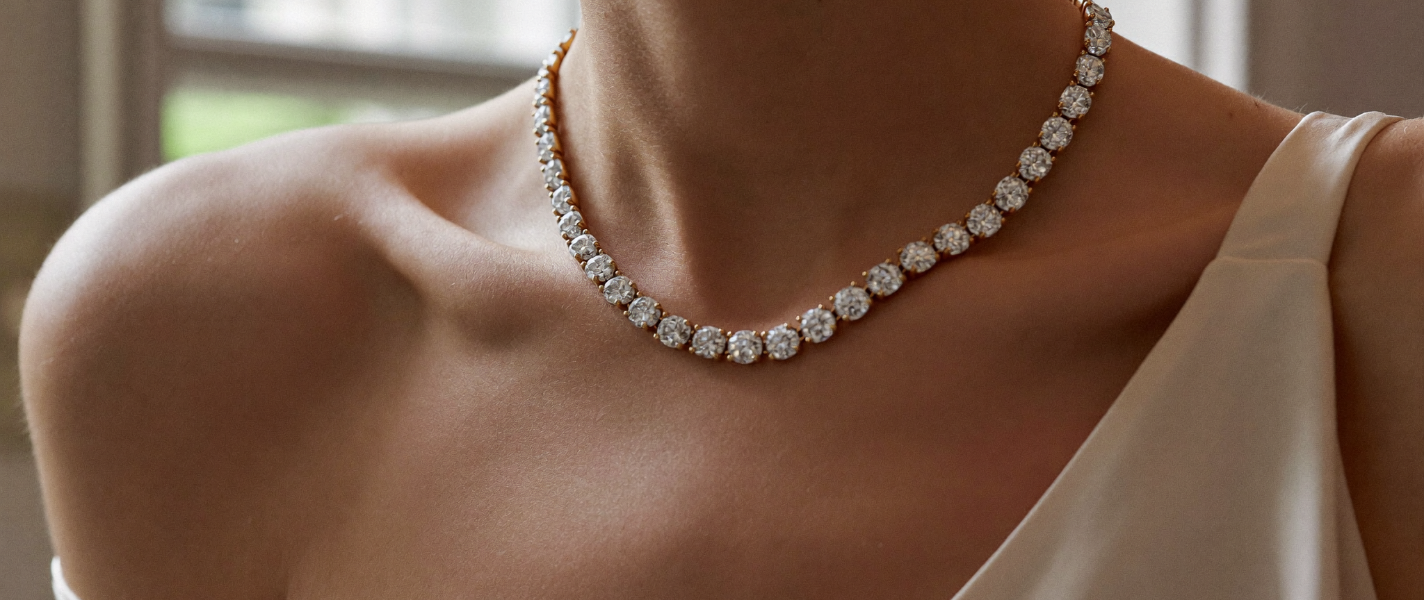 Diamond tennis necklace in gold worn across the collarbone with a cream silk slip, natural window light