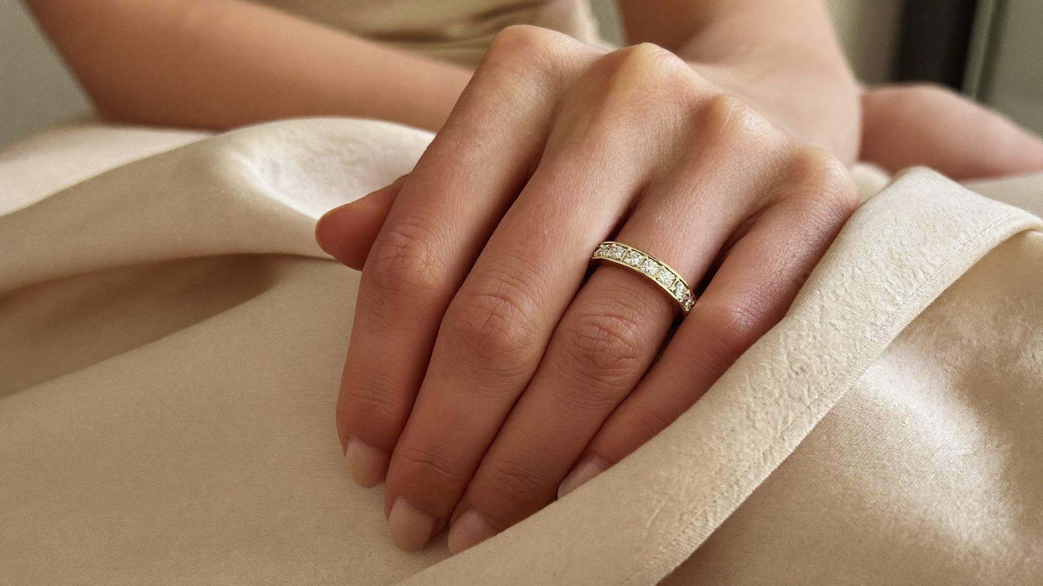 Diamond eternity band in solid yellow gold worn on a hand resting on cream silk, natural window light