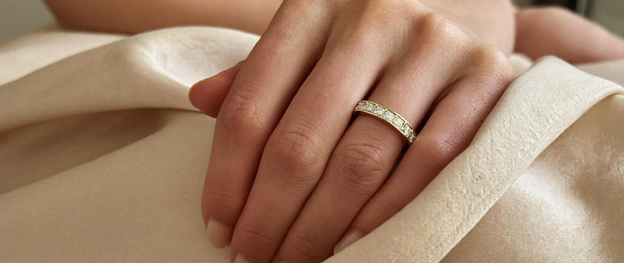Diamond eternity band in solid yellow gold worn on a hand resting on cream silk, natural window light